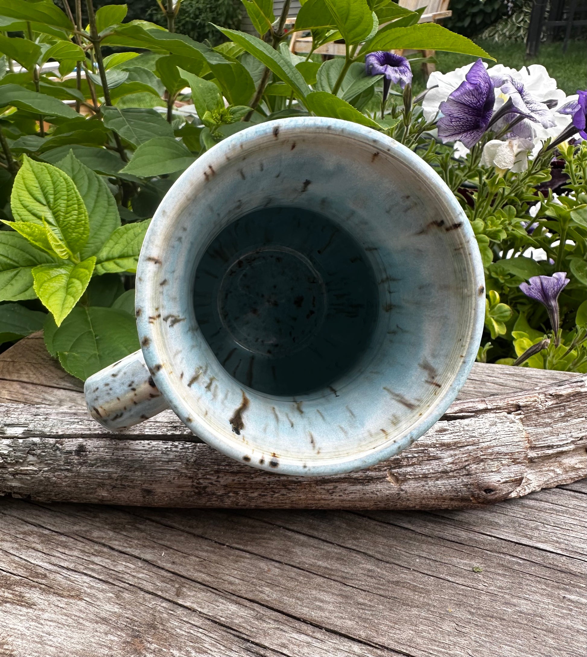 A handmade ceramic mug with a distressed light blue and white glaze, shown from a top-down perspective on a weathered wooden log with garden foliage and purple flowers in the background.