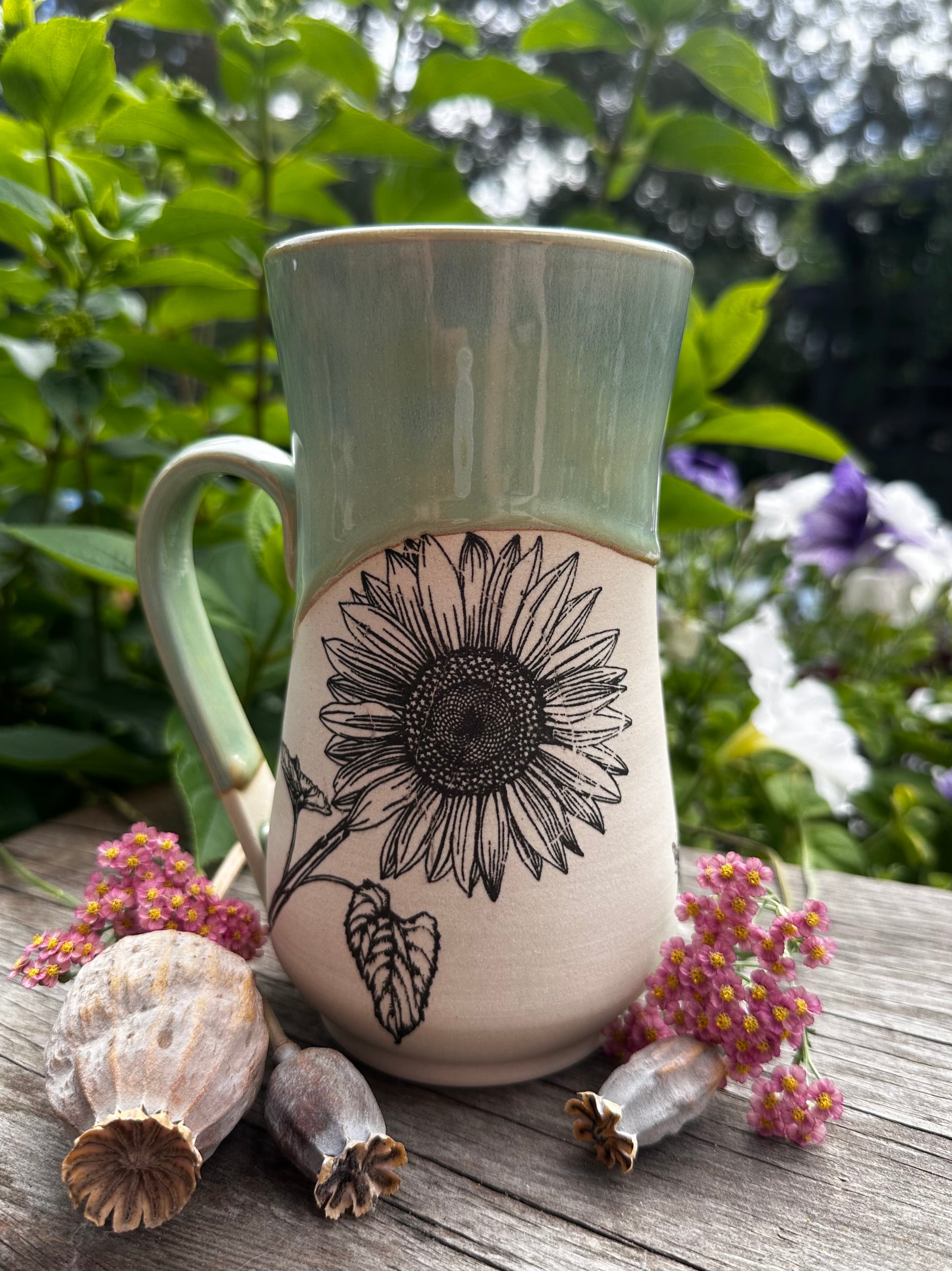 A ceramic mug with a light green glazed upper half and handle, and a cream-colored lower half featuring a black hand-drawn sunflower design. The mug is styled on a rustic wooden surface with dried poppy pods and small purple flowers, set against a soft-focus background of green garden foliage.